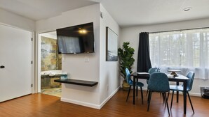 Ein modernes Wohnzimmer mit einem an der Wand befestigten Flachbildfernseher, einem Essbereich mit Tisch und Stühlen sowie einem großen Fenster mit Vorhängen.