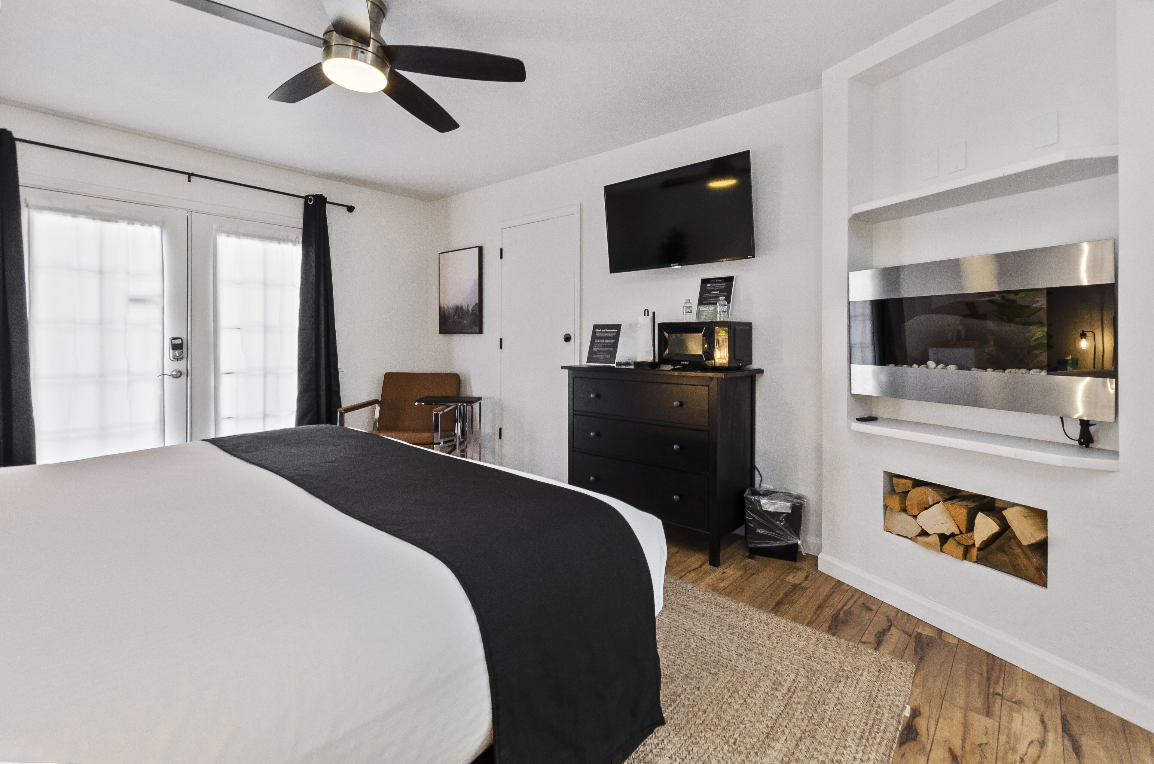 A modern bedroom with a large bed, a ceiling fan, a flat-screen TV, a fireplace, and a wooden floor.