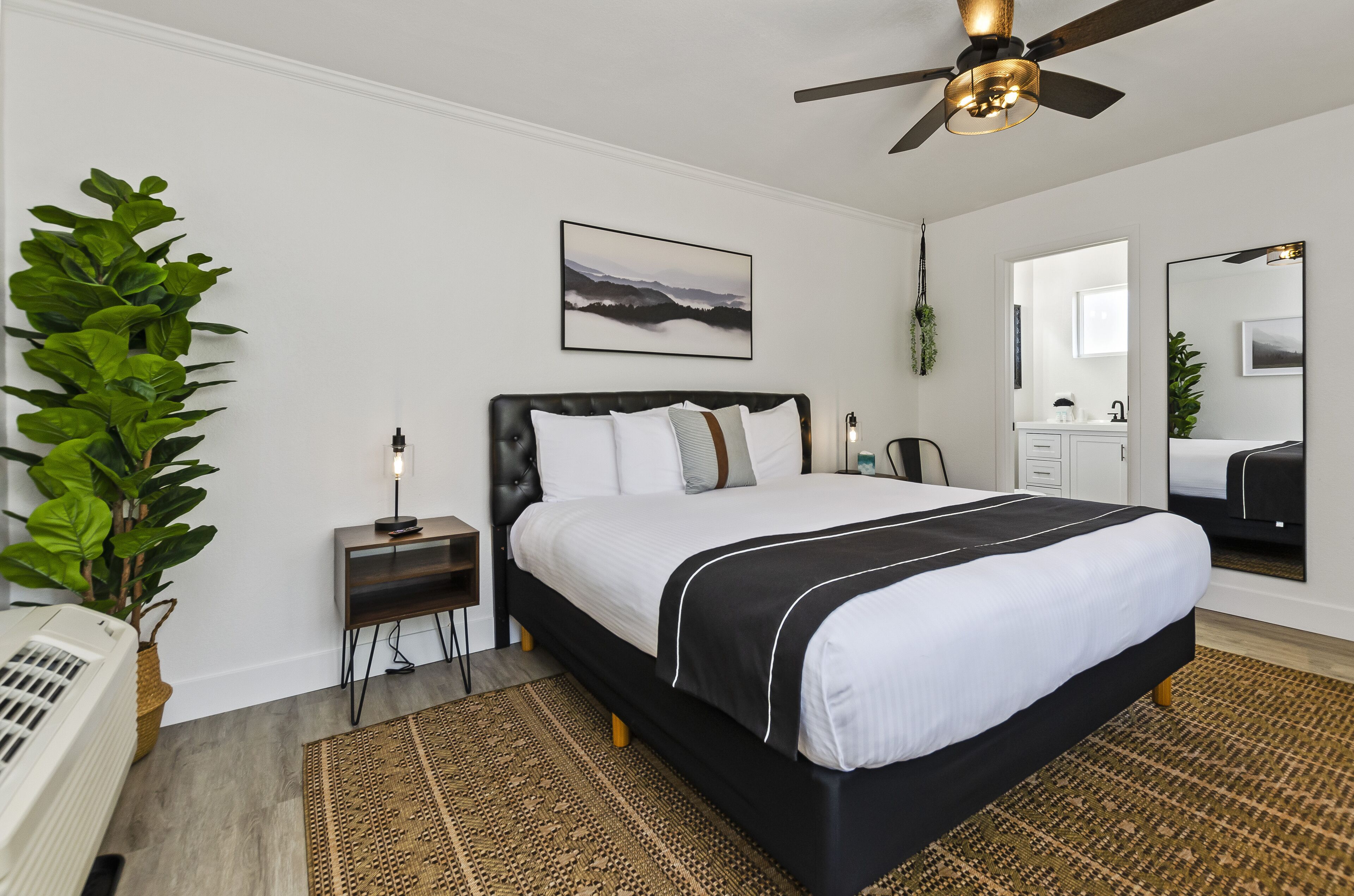 A modern bedroom with a large bed, a ceiling fan, a bedside table with a lamp, a wall-mounted picture, and a large mirror.