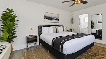 A modern bedroom with a large bed, a ceiling fan, a bedside table with a lamp, a wall-mounted picture, and a large mirror.
