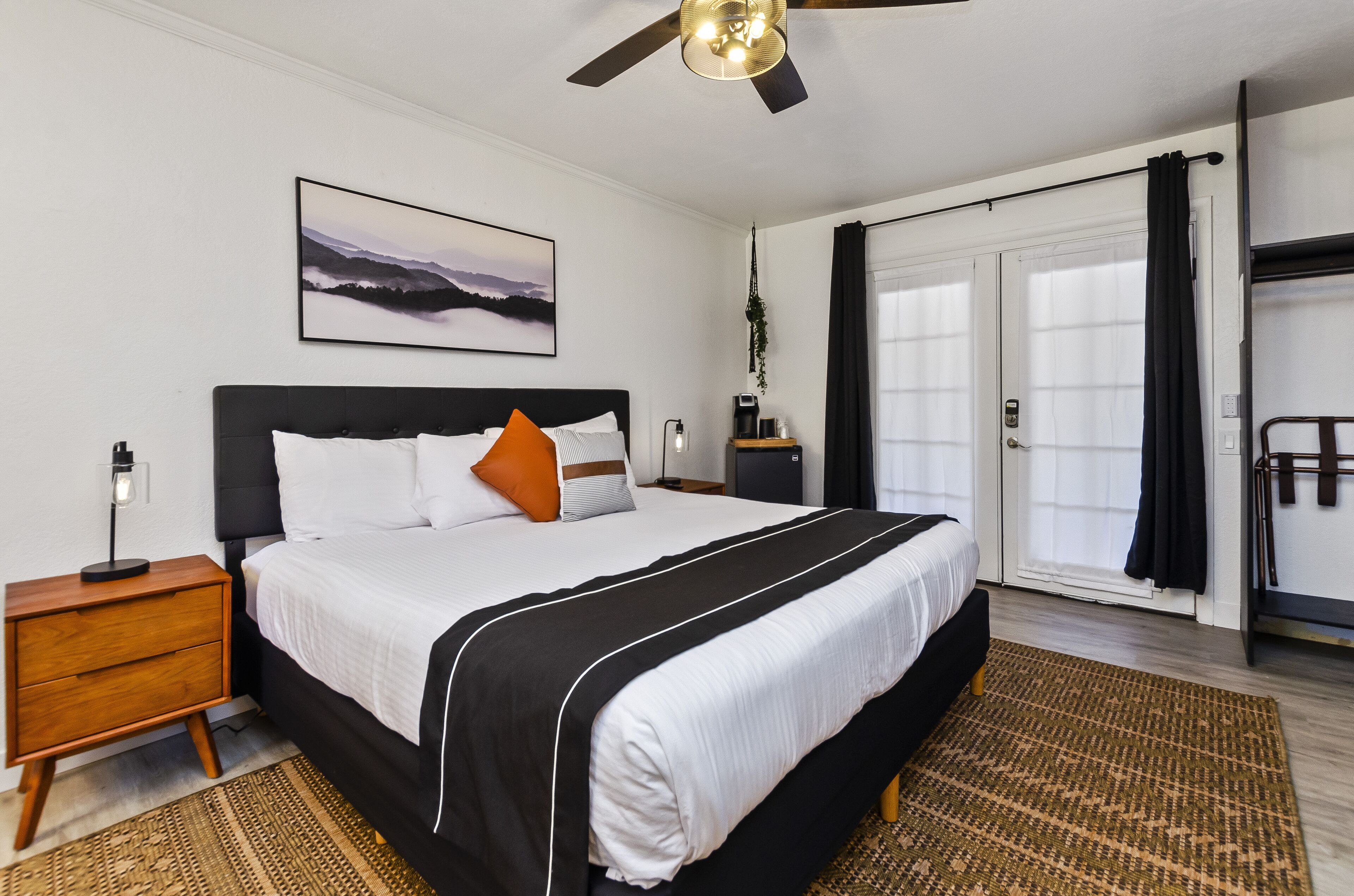 A bedroom with a large bed, a nightstand, a ceiling fan, a painting, a window with curtains, and a door.