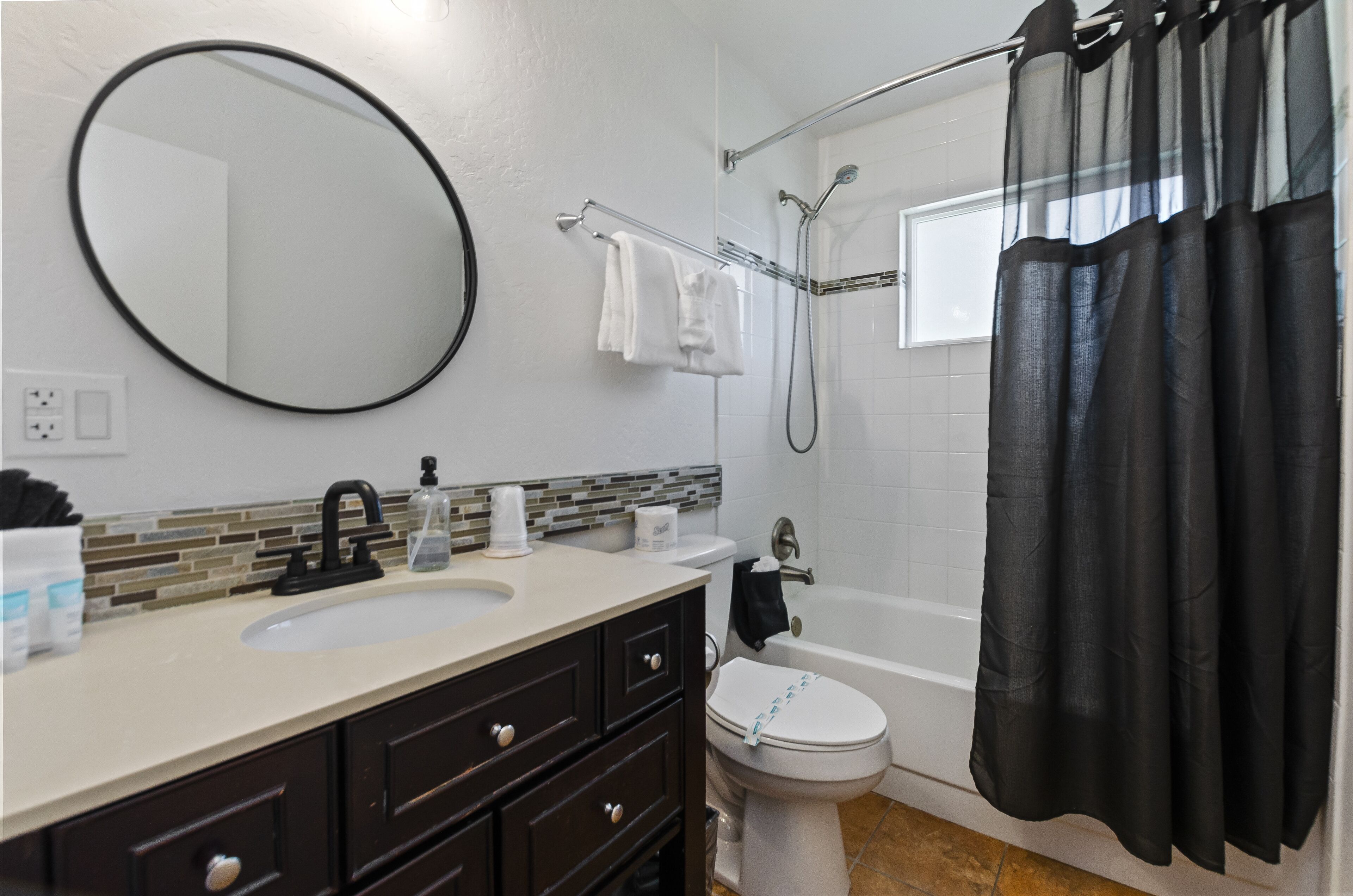 Un baño con espejo redondo, lavabo con grifo, inodoro, bañera con ducha y cortina negra.