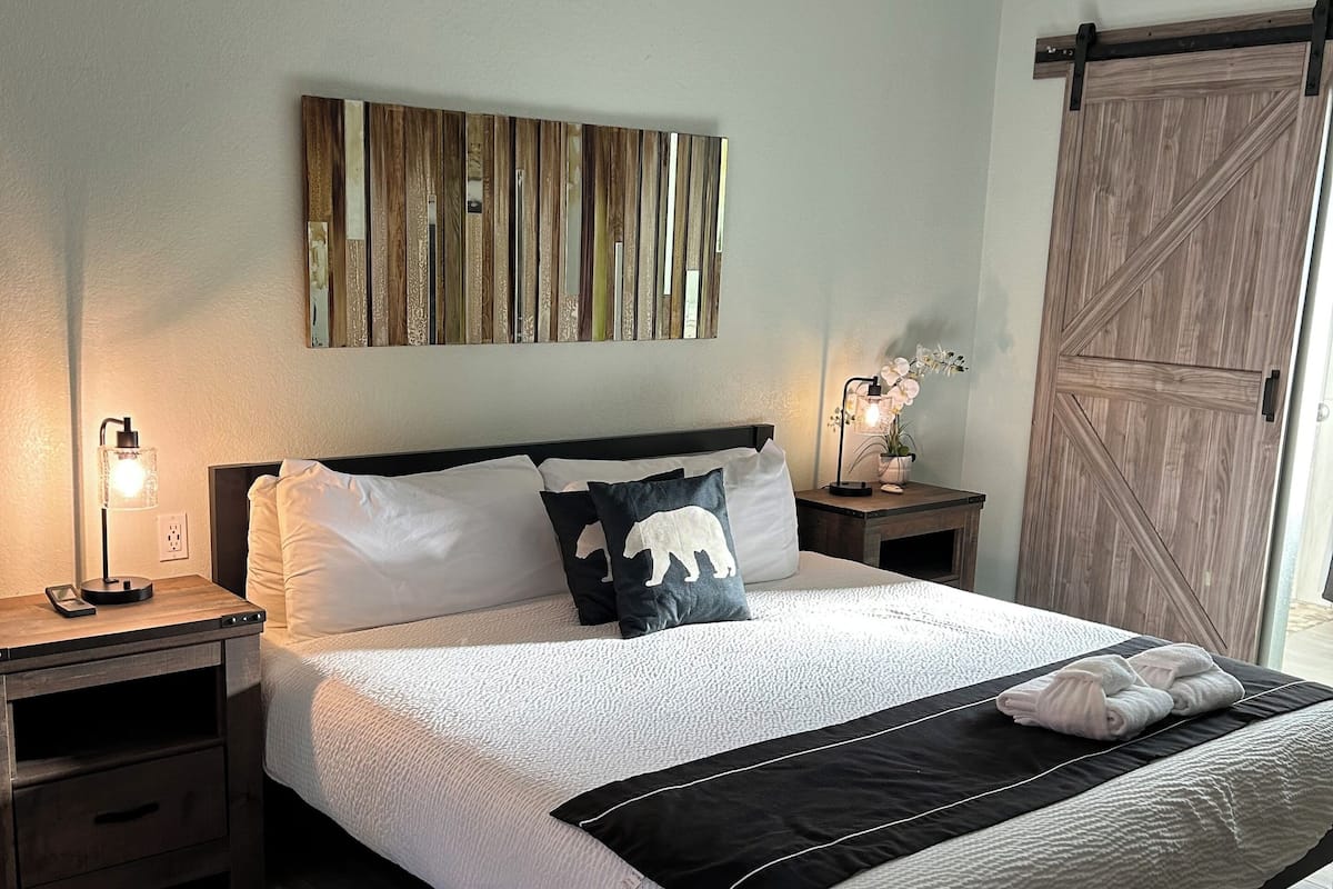 A neatly made bed with a white comforter and black accent pillows, a wooden nightstand with a lamp, a wall-mounted abstract art piece, and a wooden door.