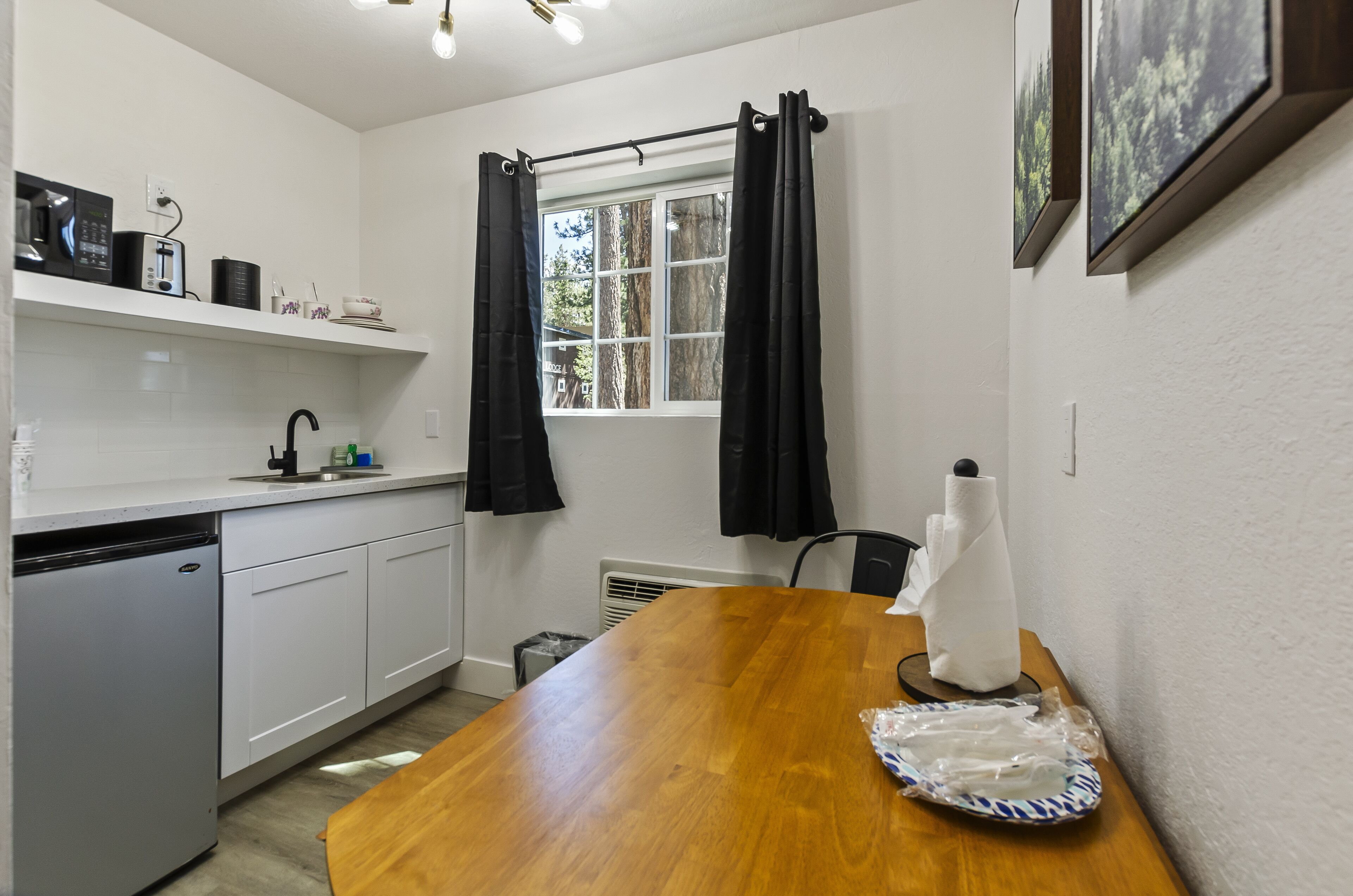 A compact kitchen with white cabinets, a wooden dining table, a microwave, a toaster, and a window with black curtains.