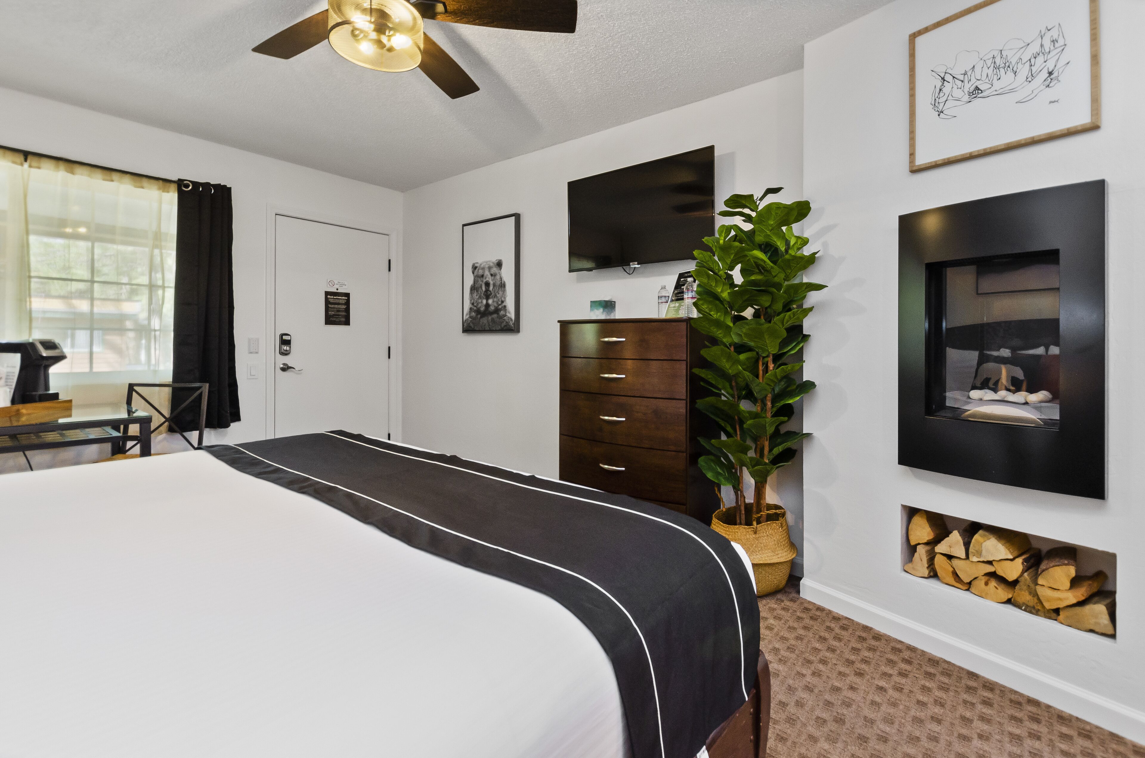 A hotel room with a large bed, a desk with a computer, a television, a plant, and a fireplace.