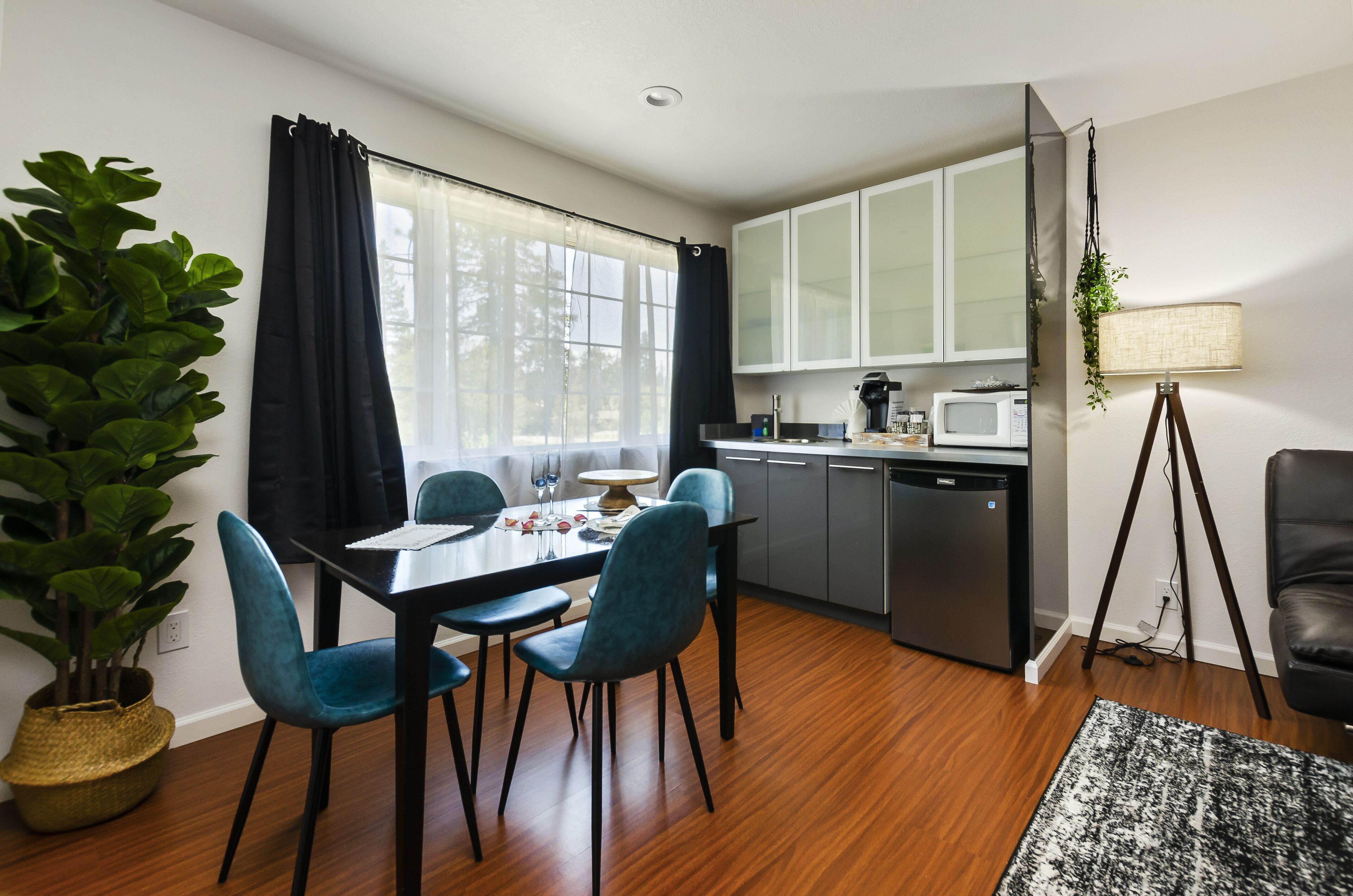 Una cocina moderna con armarios blancos, microondas y cafetera. Hay una mesa de comedor con sillas y una planta en maceta.