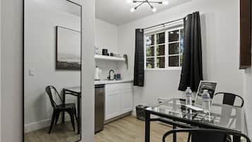 Una cocina moderna con armarios blancos, un lavavajillas de acero inoxidable y una encimera negra. Hay una mesa de comedor con sillas y una cortina negra sobre una ventana.