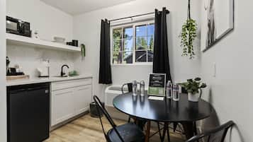 A modern kitchen with white cabinets, a black refrigerator, and a dining table with chairs. There is a window with black curtains, a hanging plant, and a framed picture on the wall.