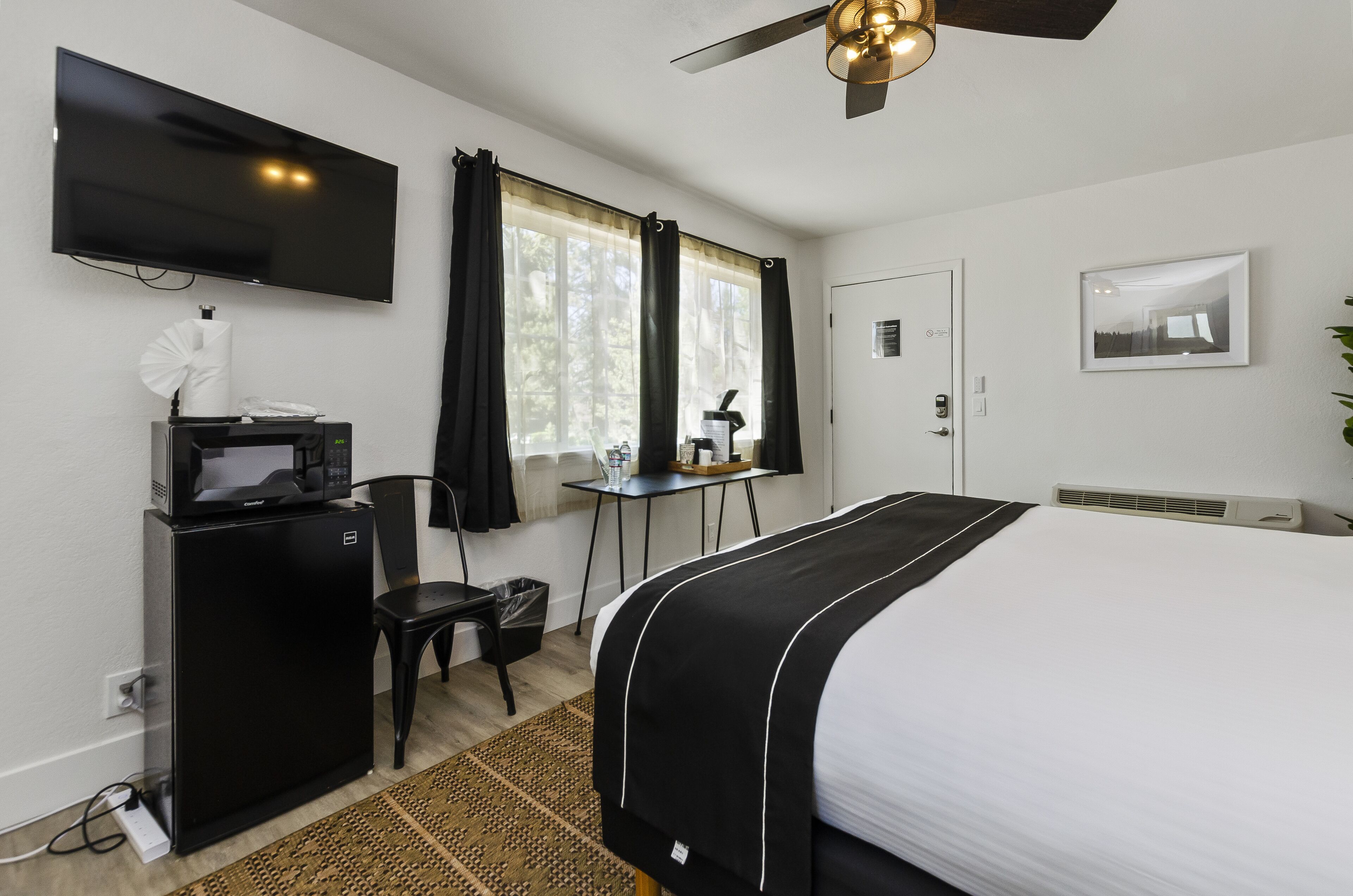 Habitación de hotel con una cama, un escritorio con microondas, una silla, televisor, ventana con cortinas y ventilador de techo.
