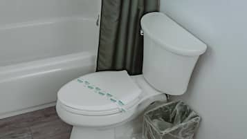 Un inodoro blanco con tapa, un cubo de basura de plástico y una bañera en un baño.