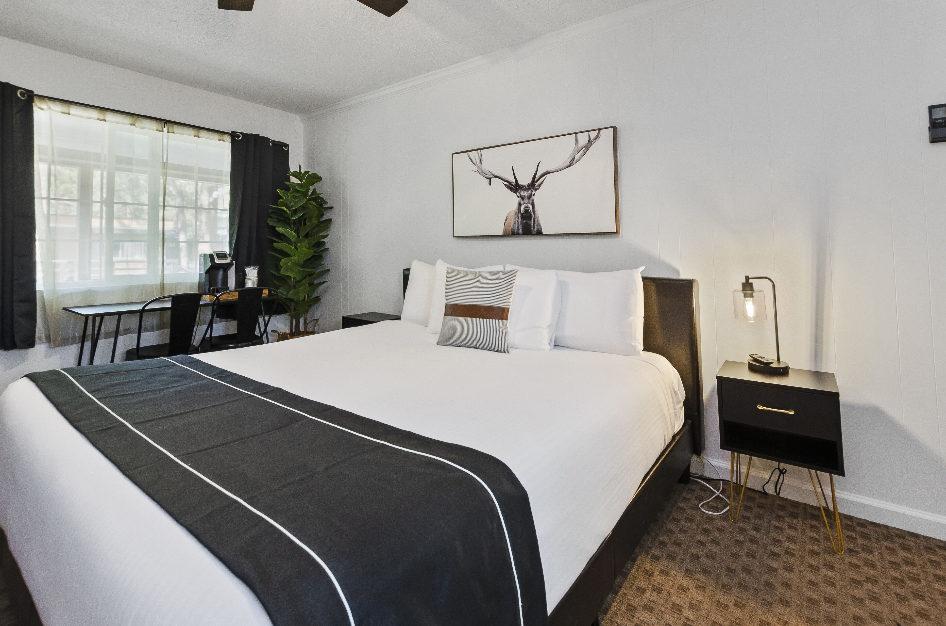 Ein Hotelzimmer mit einem großen Bett, einem Schreibtisch, einem Sessel, einer Topfpflanze, einem Nachttisch und einem Wandbild von einem Reh.