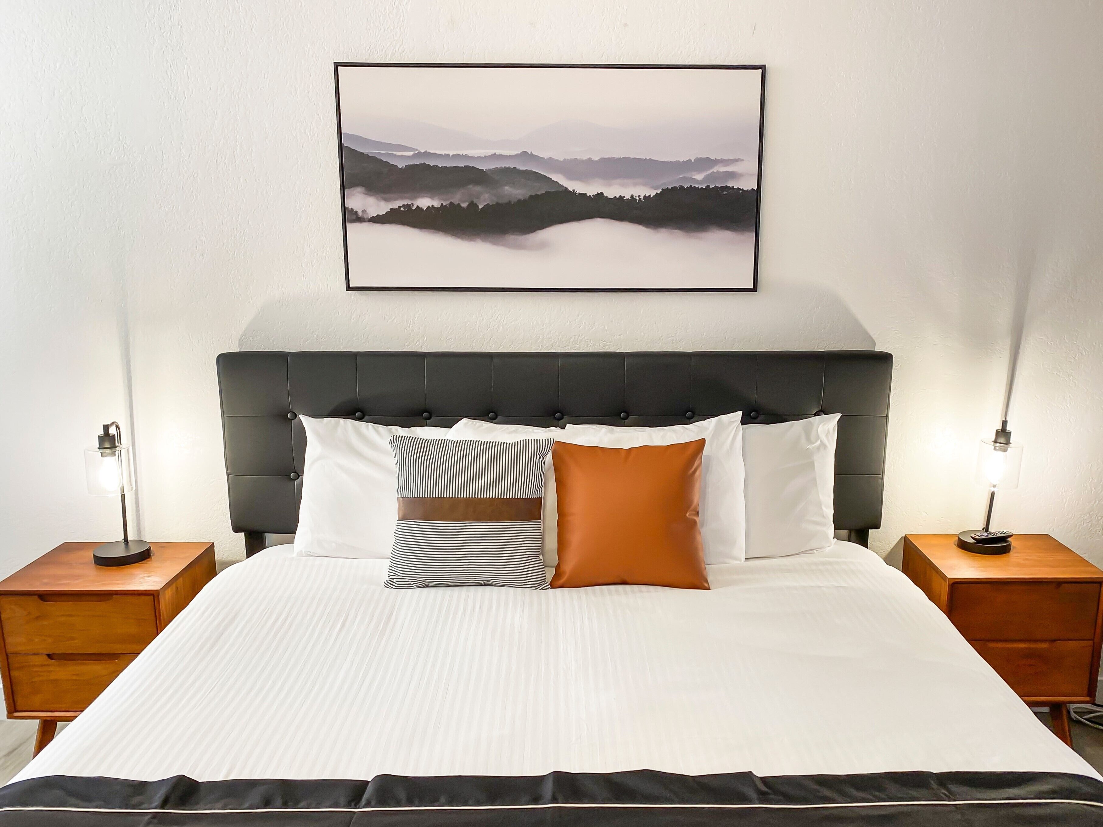 Ein ordentlich gemachtes Bett mit einem gestreiften Kissen, einem einfarbig orangefarbenen Kissen und einem weißen Kissen. An der Wand über dem Bett hängt ein gerahmtes Landschaftsbild. Zwei Nachttische stehen an beiden Seiten des Bettes, jeweils mit einer Lampe.