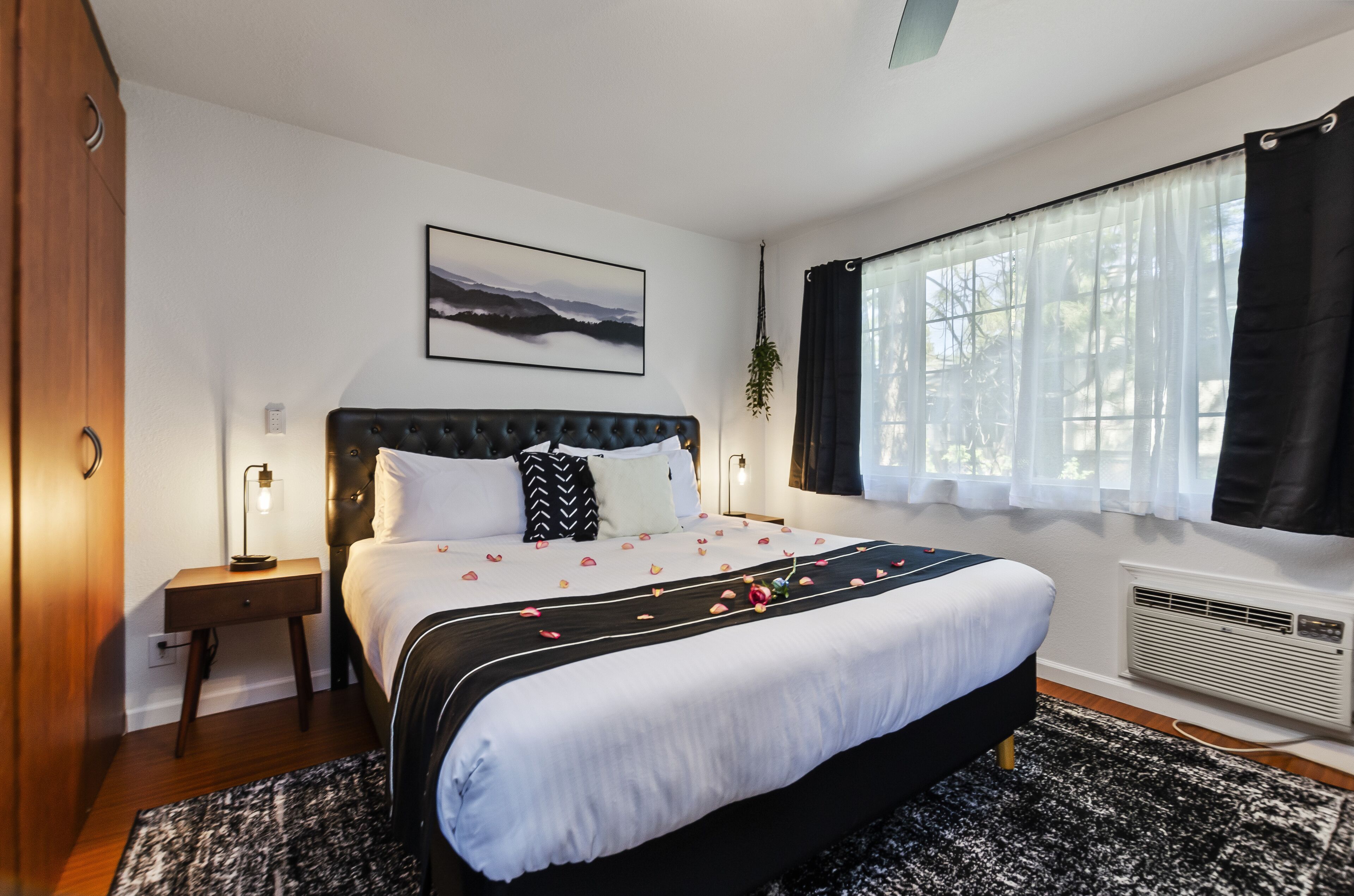 Une chambre avec un grand lit, une table de chevet, une lampe fixée au mur, une fenêtre avec des rideaux et un tableau encadré accroché au mur.
