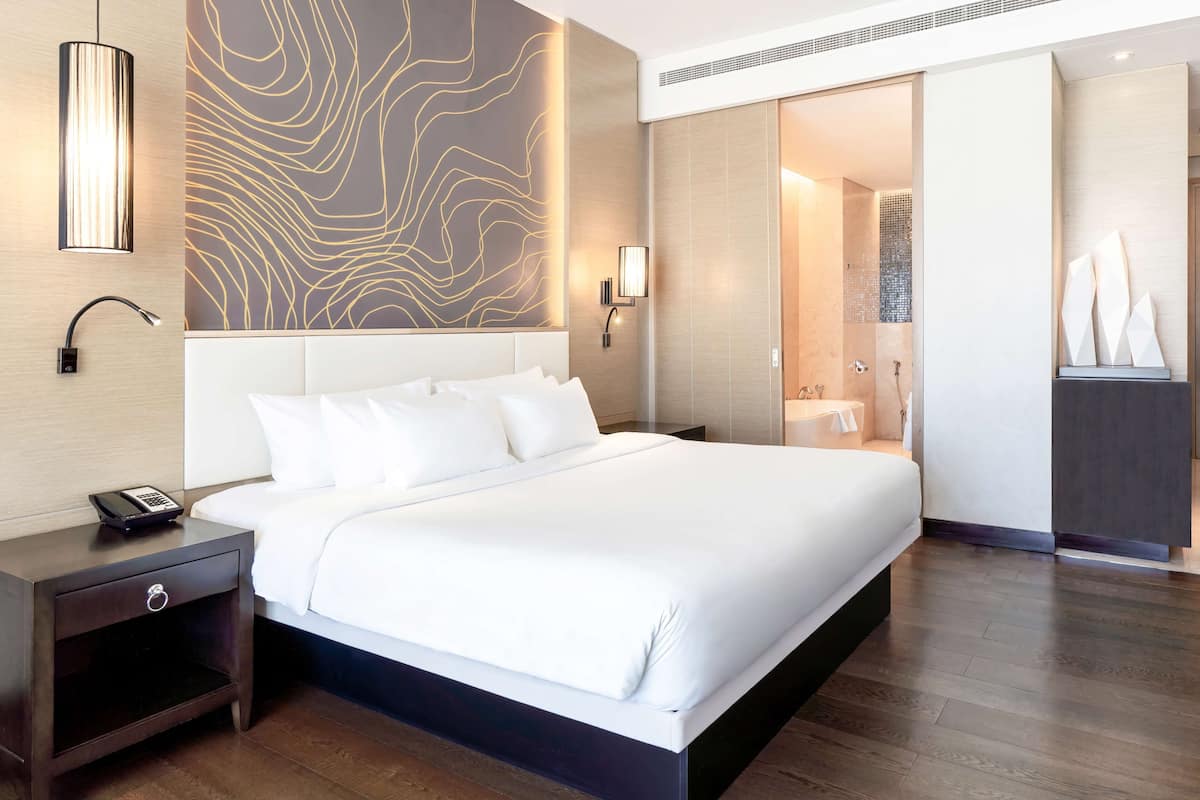 A modern hotel room with a large bed, a nightstand with a phone, a wall-mounted light, and a bathroom visible through an open door.