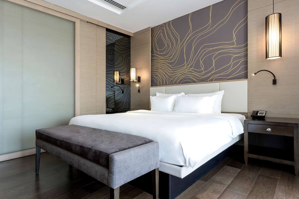A modern hotel room with a large bed, a bench, a nightstand, and wall-mounted lighting.
