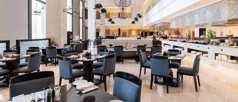 A spacious hotel lobby with multiple dining areas, modern lighting fixtures, and a large kitchen visible in the background.
