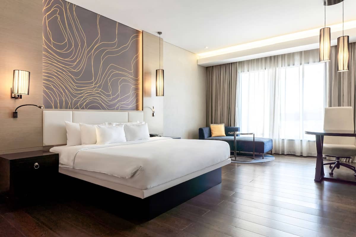 A modern hotel room with a large bed, a desk, and a chair. There is a wall with a light design, a window with curtains, and a lamp on the nightstand.