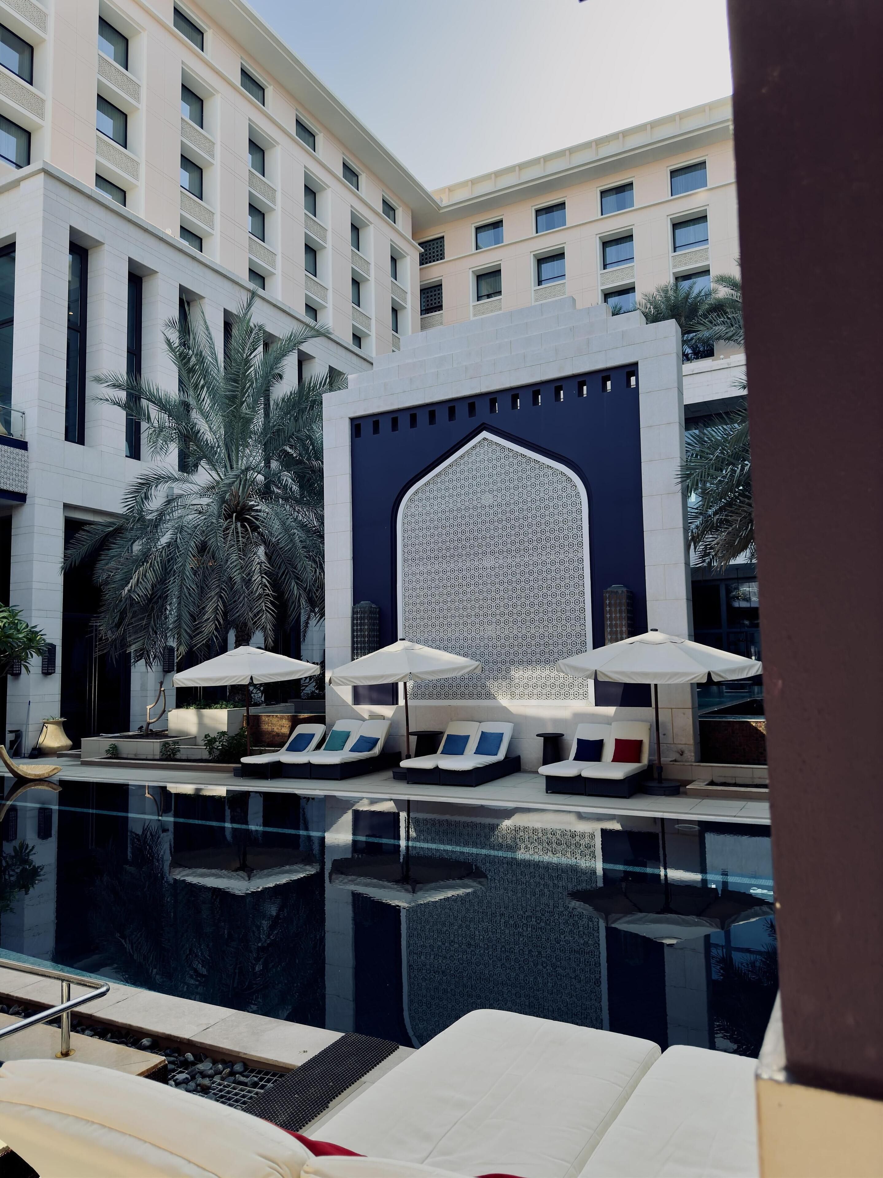 A modern hotel pool area with lounge chairs and umbrellas, surrounded by palm trees and a large decorative wall.