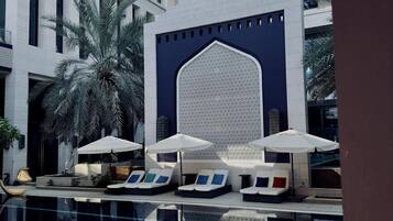 A modern hotel pool area with lounge chairs and umbrellas, surrounded by palm trees and a large decorative wall.