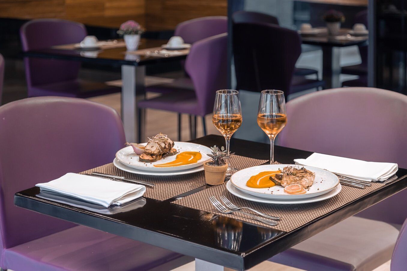 Una mesa de comedor con platos de comida, dos copas de vino y una pequeña planta.