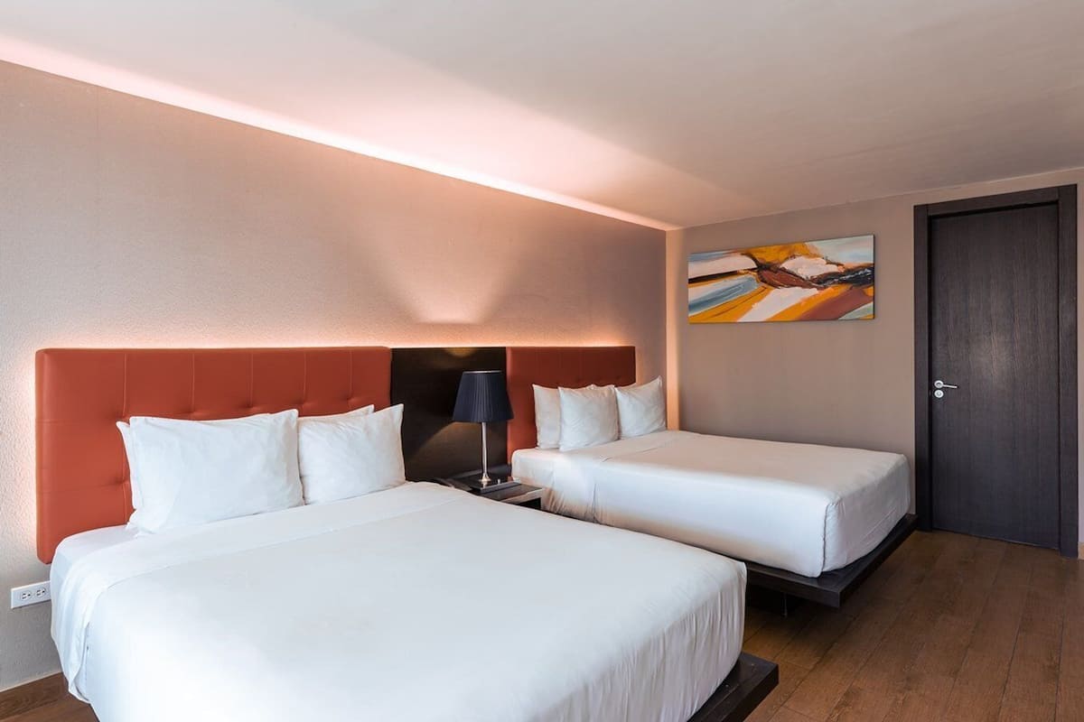 A hotel room with two beds, a nightstand, a lamp, a painting, and a door.