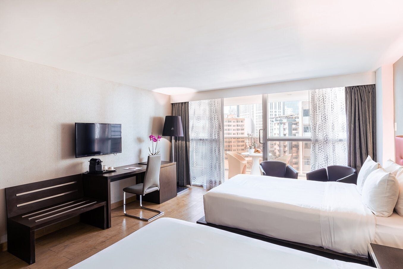 Une chambre d’hôtel avec un lit, un bureau doté d’une télévision, une chaise et une vue sur la ville grâce à de grandes fenêtres.