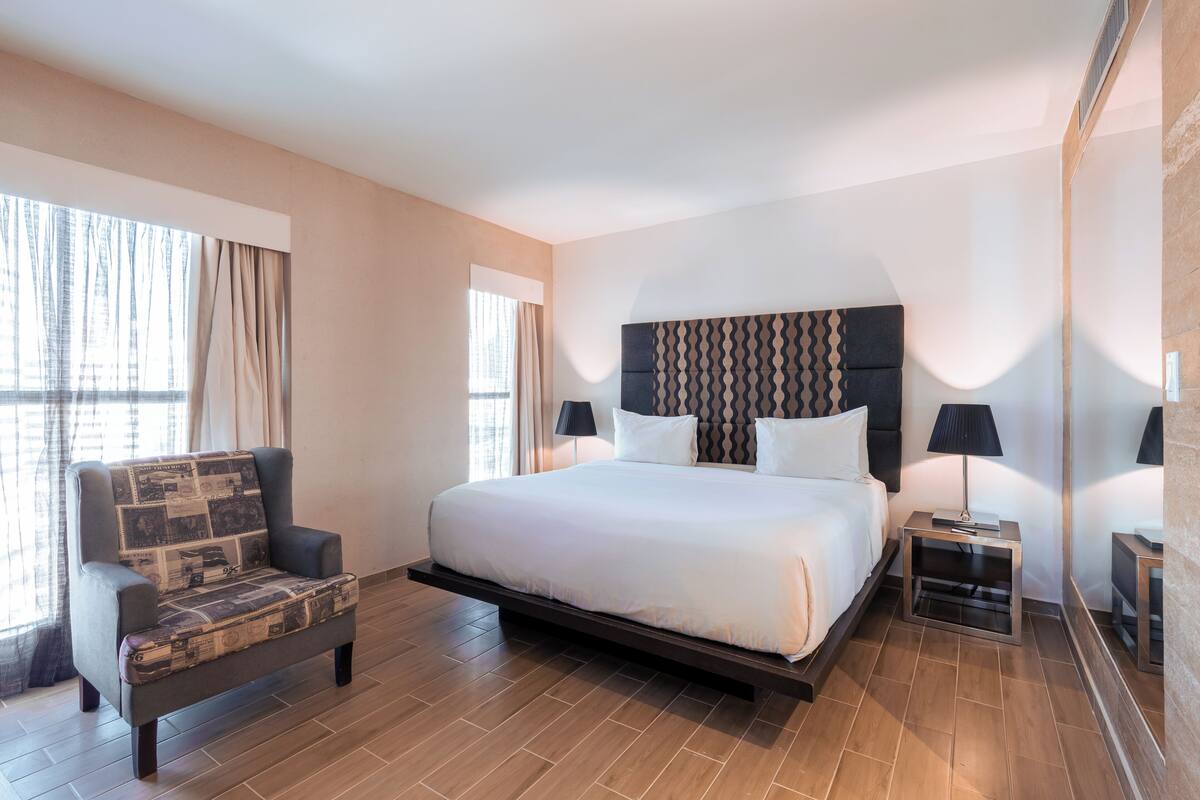 A modern hotel room with a large bed, a sofa, two bedside tables with lamps, and a large window with curtains.