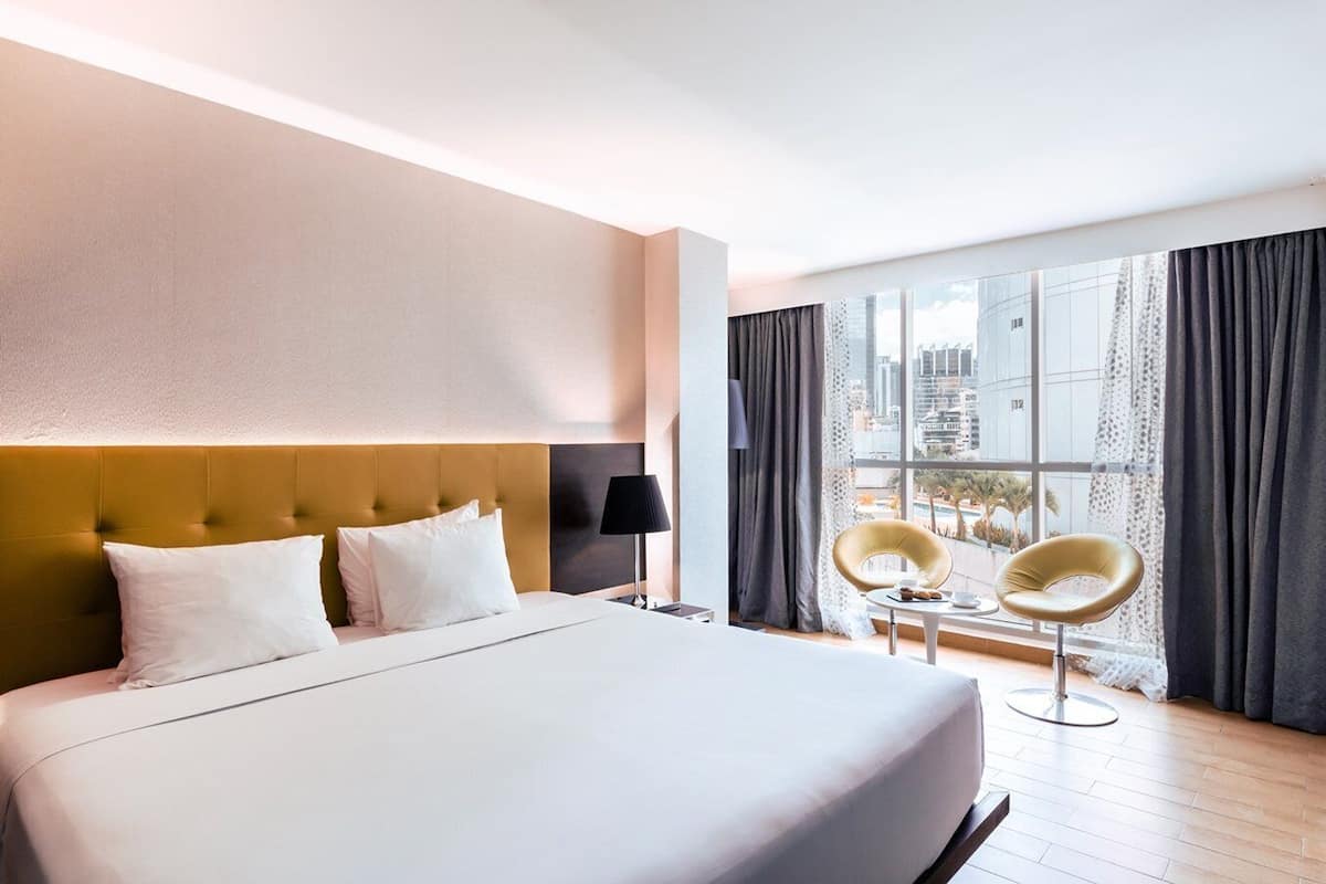 A hotel room with a large bed, two chairs, a small table, and a view of the city through large windows.