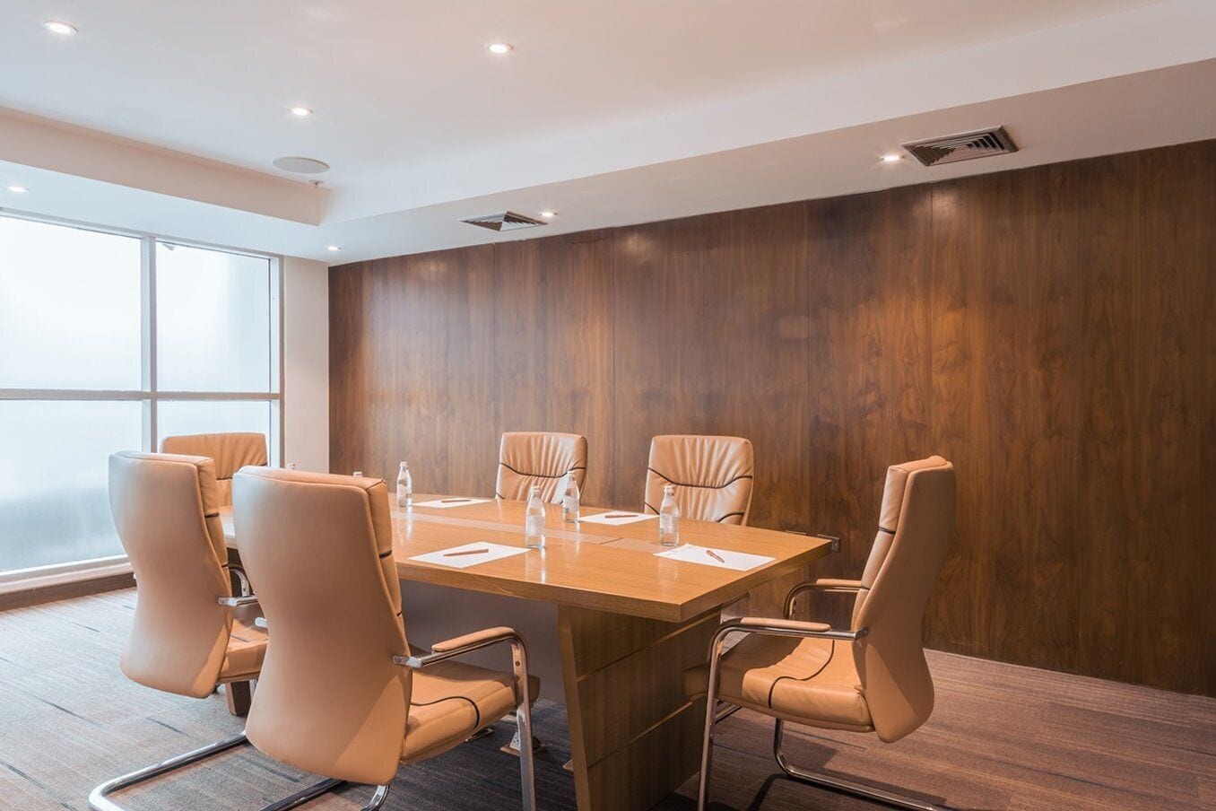 Une salle de conférence avec une table en bois, six chaises beiges et une grande fenêtre.