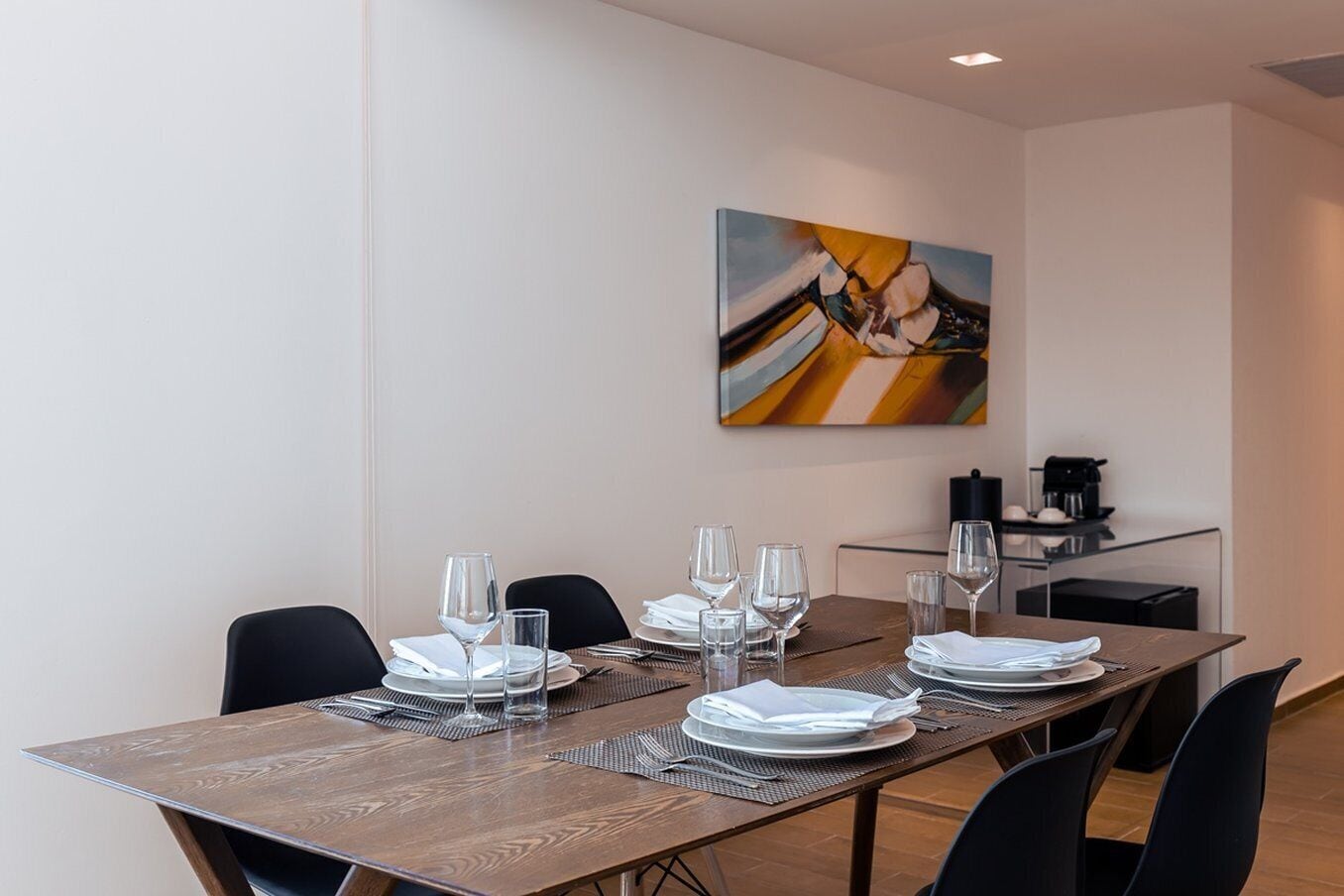 A dining area with a long table set for four, equipped with plates, glasses, and cutlery. A coffee machine is visible in the background.