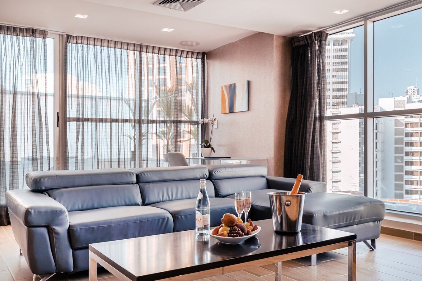 Una sala de estar moderna con un sofá gris, una mesa de centro y vistas de la ciudad a través de amplias ventanas.