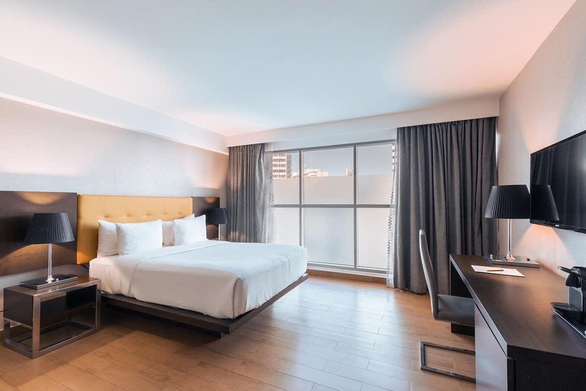A modern hotel room with a large bed, a desk with a flat-screen TV, and a large window with curtains.