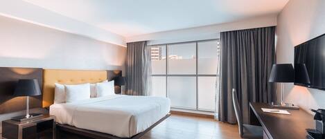 A modern hotel room with a large bed, a desk with a flat-screen TV, and a large window with curtains.