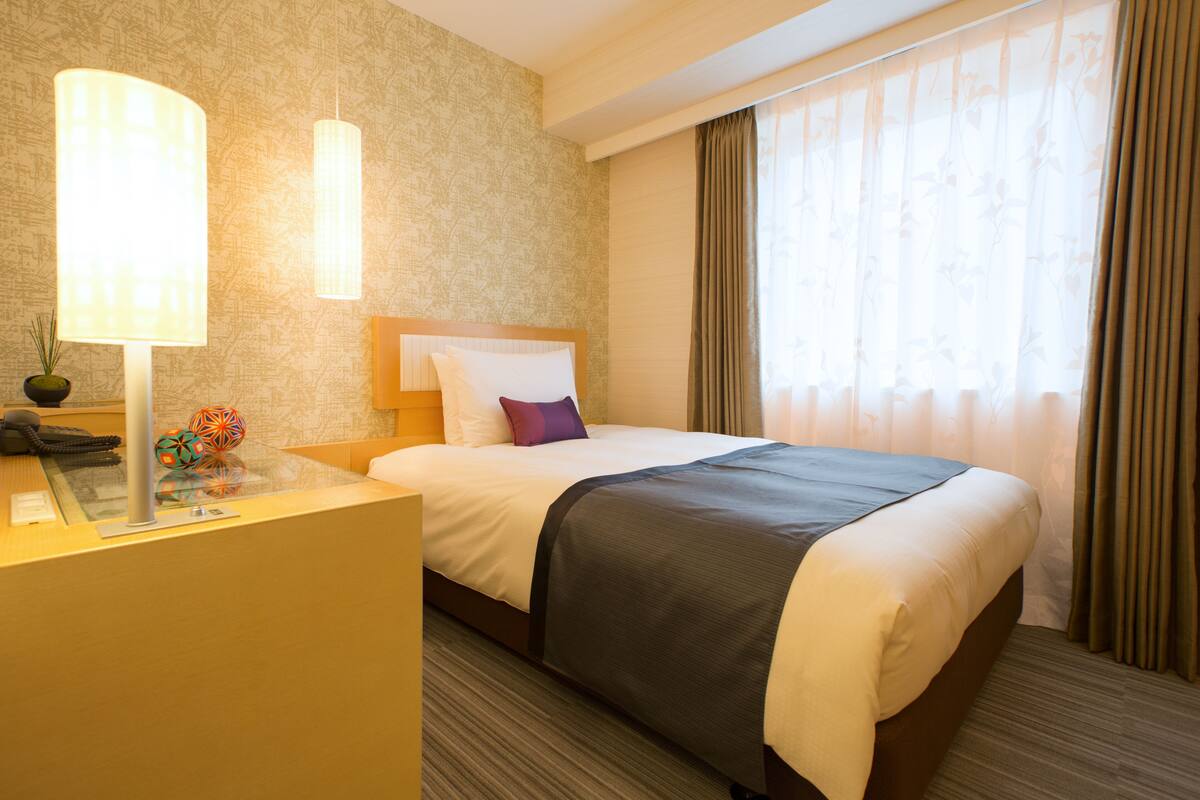 A hotel room with a bed, a desk with a lamp, and a window with curtains.