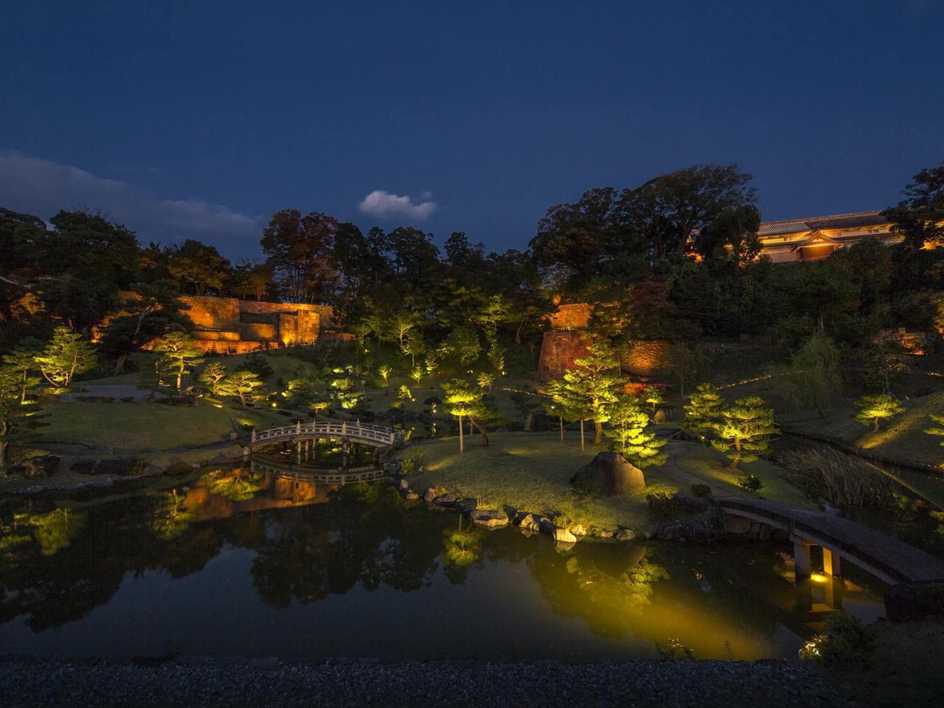 A serene night scene of a traditional Japanese garden with a bridge, pond, and illuminated pathways.