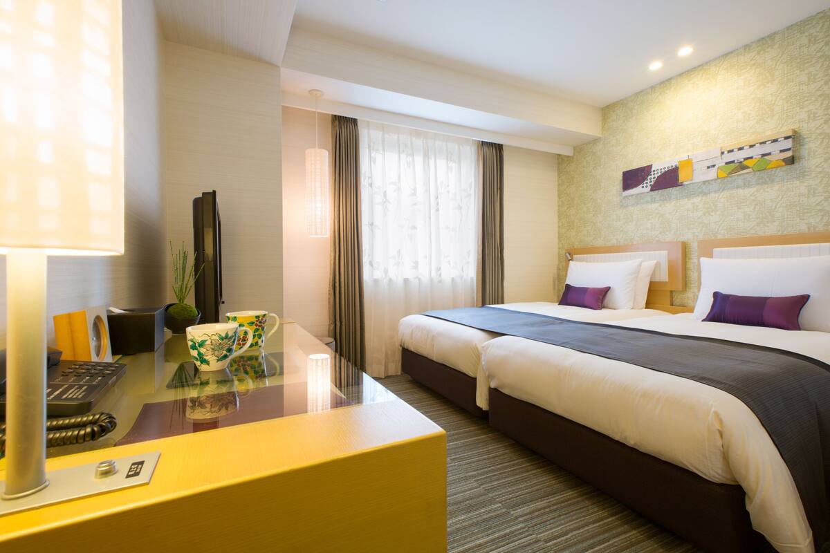 A hotel room with a large bed, a desk with a computer, a lamp, and a window with curtains.