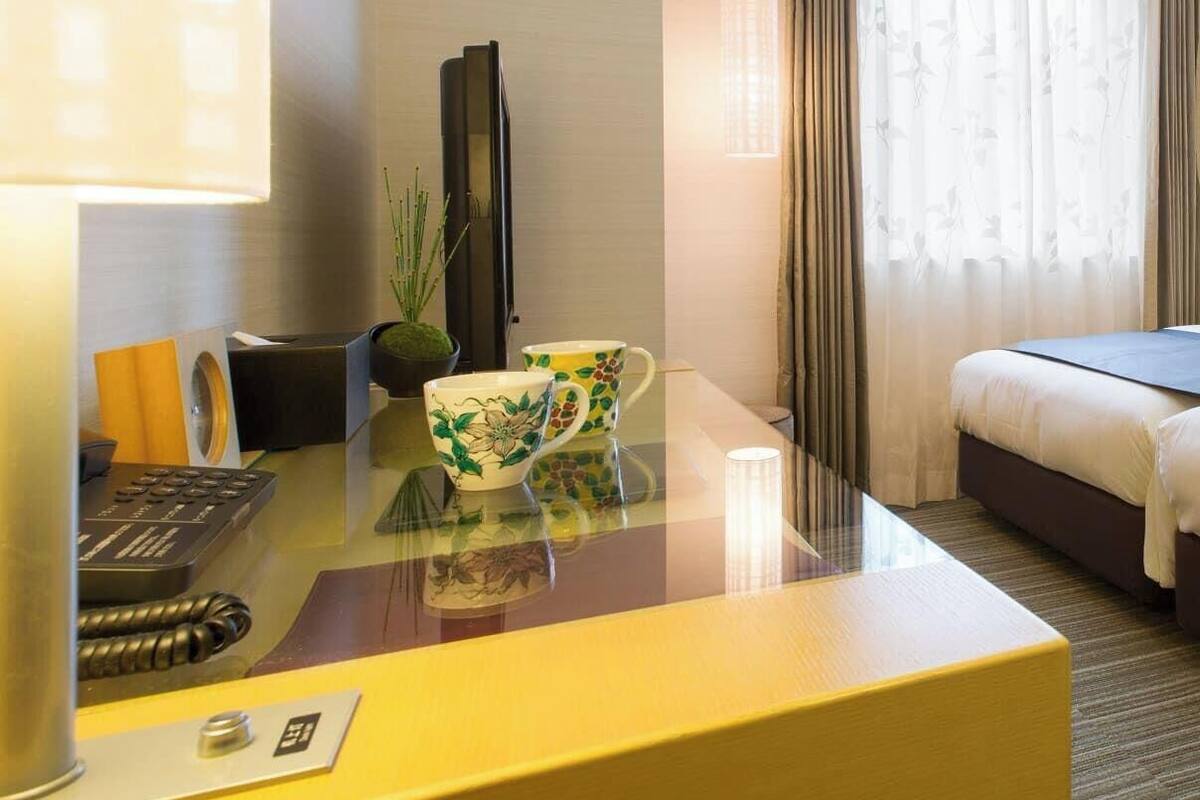 A hotel room with a bed, a desk with a lamp, a telephone, and a plant.