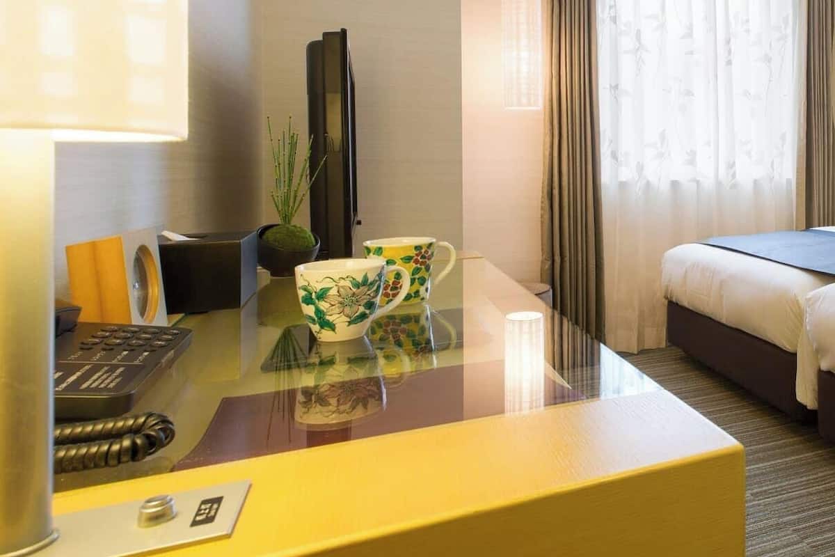 A hotel room with a bed, a desk with a lamp, a telephone, and a plant.