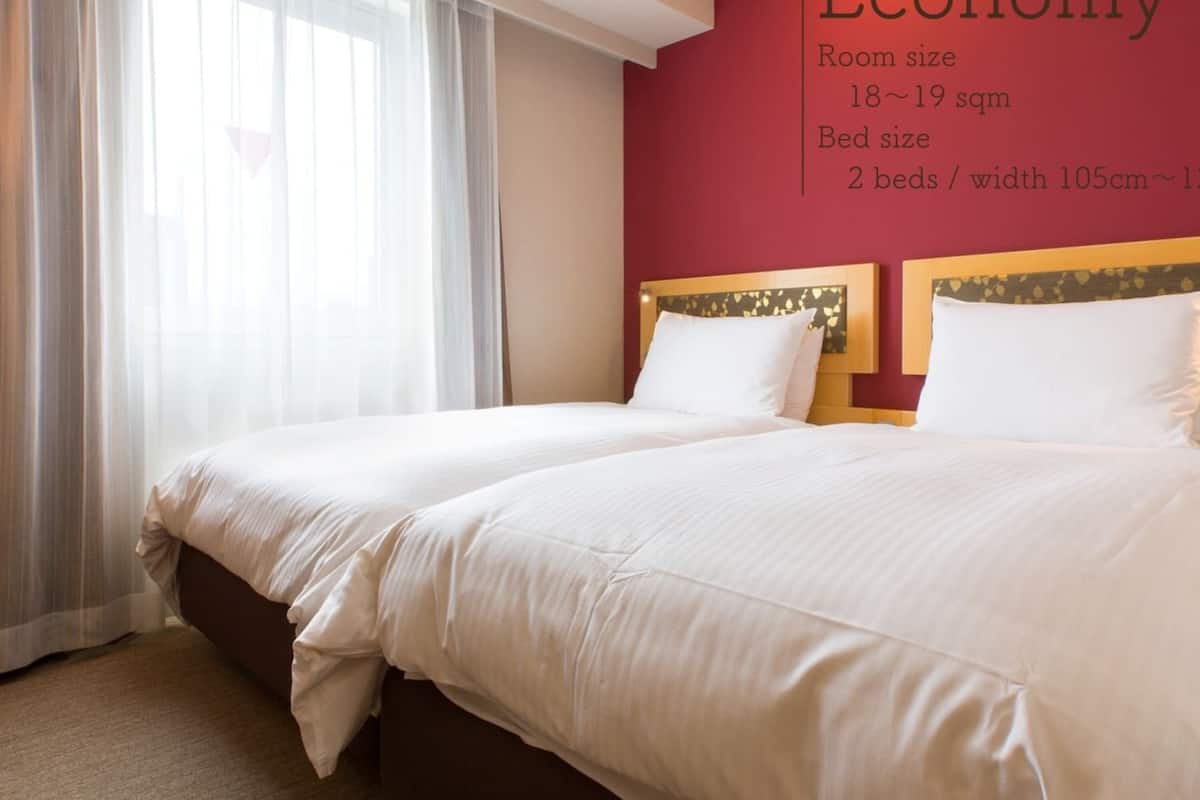 A hotel room with two beds, a red accent wall, and a large window with sheer curtains.