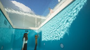 Vista subaquática de uma piscina com água azul cristalina, uma escada e uma pessoa em pé na borda.