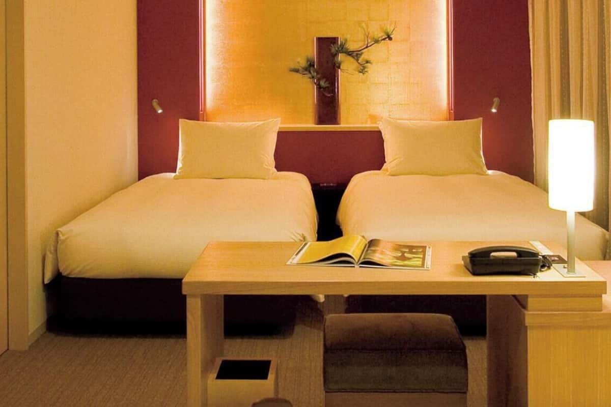 A hotel room with two beds, a bedside table with a book and a phone, a small table with a lamp, and a TV on the left.