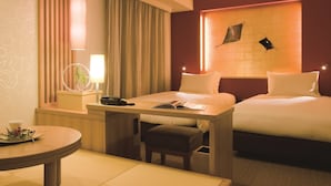 Quarto de hotel com duas camas, uma pequena escrivaninha com luminária, uma cadeira e uma mesa redonda com uma planta.