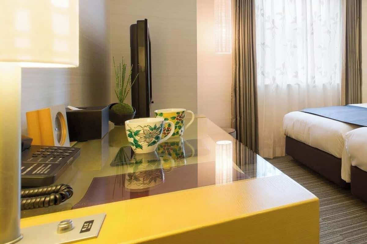 A hotel room with a bed, a desk with a lamp, a plant, and a window with curtains.
