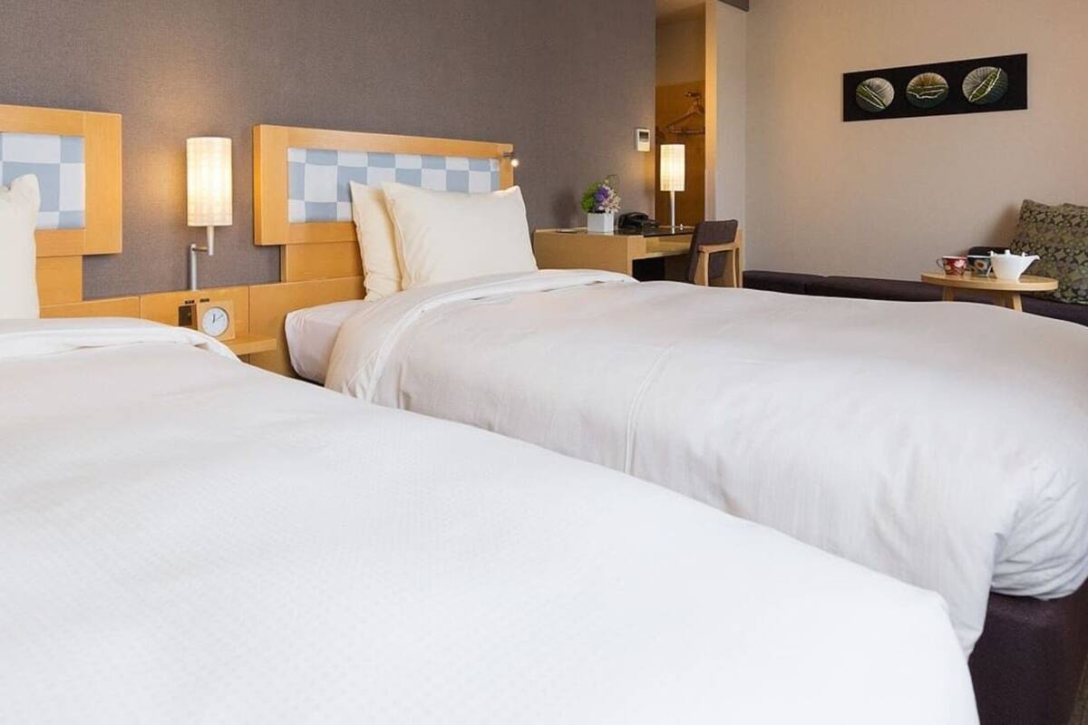 A hotel room with two beds, a desk, a chair, and a small table with a lamp.