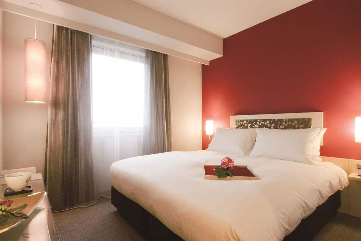 A hotel room with a large bed, a bedside table with a lamp, a window with curtains, and a red accent wall.