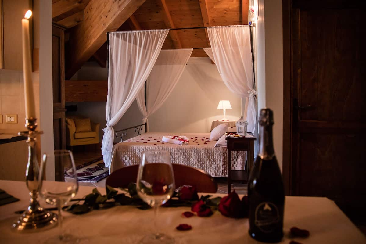 Ein gemütliches Schlafzimmer mit einem Bett, einem Nachttisch mit einer Flasche, sowie einem Tisch mit Weingläsern und Rosen.