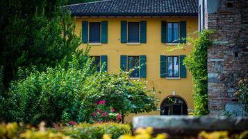 A yellow building with a tiled roof and green shutters, surrounded by lush greenery and a stone wall.