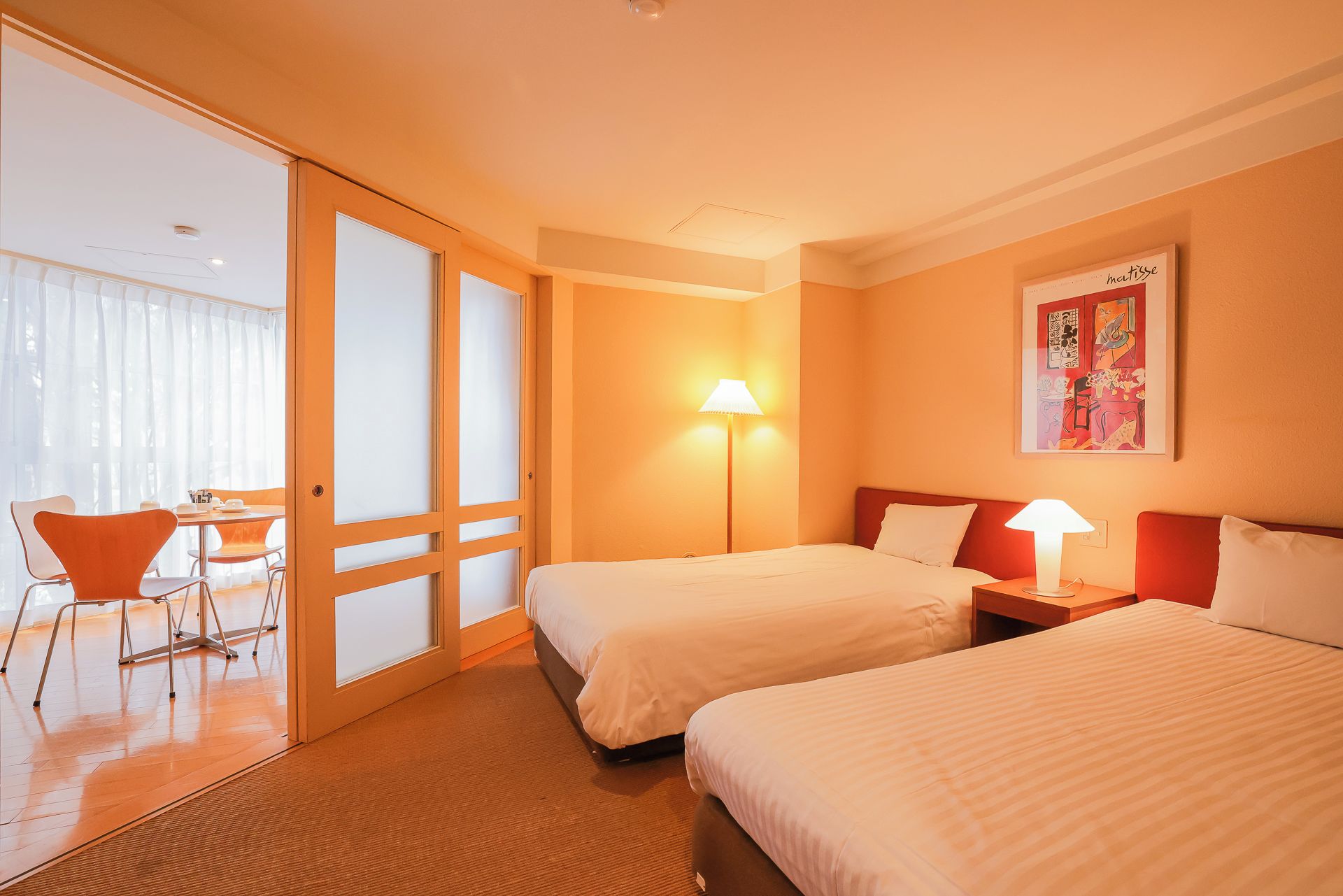 Une chambre d’hôtel avec deux lits, un coin repas comprenant une table et des chaises, et une grande fenêtre avec des rideaux légers.