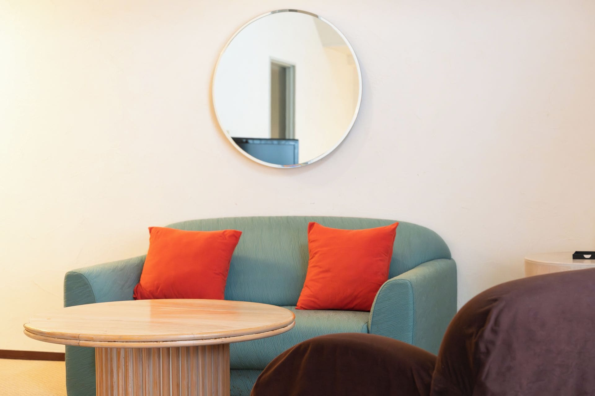 Una habitación con una mesa redonda de madera, un sofá azul turquesa con dos cojines naranjas y un espejo redondo en la pared.