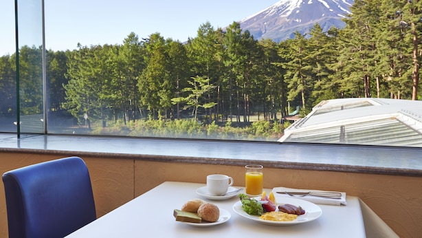 A breakfast table with a cup, juice, and plates of food, overlooking a forest and a mountain. - Fuji Premium Resort (Fujikawaguchiko)